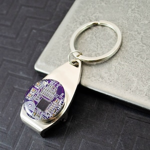 Puede incluir: Un abridor de botellas de llavero plateado con un diseño redondo de placa de circuito púrpura y plateado.