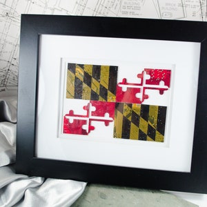 Può includere: Un'opera d'arte incorniciata che presenta la bandiera dello stato del Maryland, realizzata con circuiti stampati riciclati. La bandiera è esposta in una cornice nera con un passe-partout bianco.