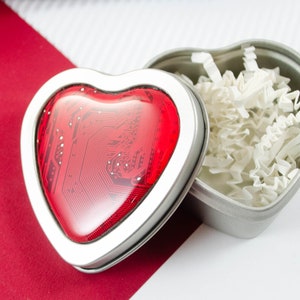 Peut inclure: Une boîte en métal argenté en forme de cœur avec un couvercle en forme de cœur rouge avec un motif de circuit imprimé. La boîte ouverte révèle du papier froissé blanc. Fond rouge et blanc.