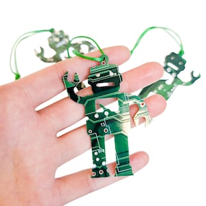 May include: A robot-shaped ornament crafted from a green printed circuit board featuring visible copper traces, solder pads, and the text T3 and R178. The ornament includes a green ribbon loop for hanging, showcasing a technical aesthetic.