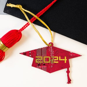 May include: A red graduation cap ornament with the year 2024 in gold. The cap is made from a circuit board design. A red tassel and black graduation cap are in the background.