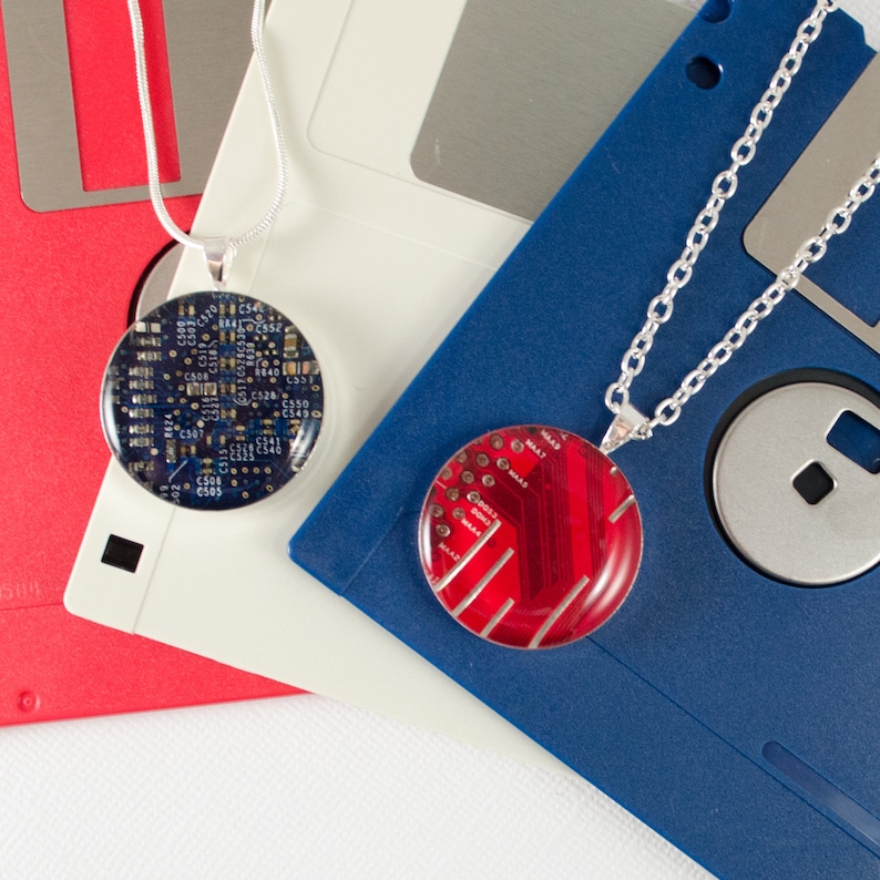 Puede incluir: Dos collares con colgantes redondos hechos de placas de circuito. Un colgante es azul y el otro es rojo. Los collares se muestran sobre disquetes azules y blancos.