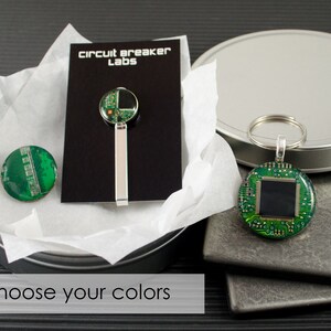Puede incluir: Un juego de dos llaveros de circuito verde, uno con un cuadrado negro en el centro y el otro con un clip de corbata plateado. Los llaveros y el clip de corbata están empaquetados en una lata plateada con un forro de papel de seda blanco. La lata tiene una etiqueta negra que dice "circuit breaker labs".
