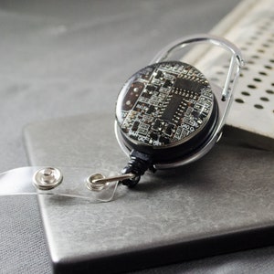 以下が含まれることがあります： 回路基板のデザインが施された、黒とシルバーのリトラクタブルバッジリール。リールは、スナップ留め具付きの透明なプラスチックストラップと、シルバーのカラビナクリップを備えています。デザインは、詳細な回路基板パターンが特徴です。