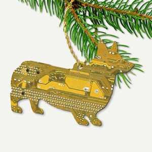 May include: A corgi-shaped Christmas ornament made from a circuit board. The ornament is gold and brown, with a detailed pattern of electronic components. It hangs from a gold string and is set against a backdrop of green pine needles.