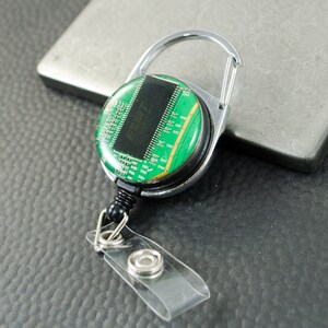 Puede incluir: Un portagafete retráctil verde y plateado con un diseño de placa de circuito. El portagafete tiene un clip giratorio plateado y una correa de plástico transparente.