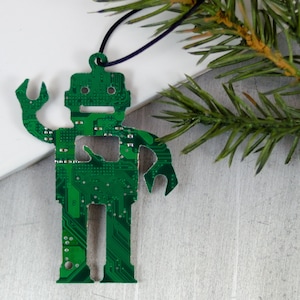 May include: A green robot-shaped Christmas ornament made from a circuit board. The ornament has a black string for hanging and is displayed with a sprig of evergreen. The robot has a wrench-like arm.