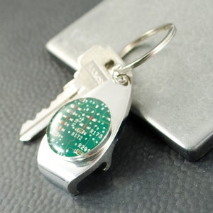 May include: A silver bottle opener keyring with a round green circuit board design. The keyring has a silver ring and is attached to a silver key.