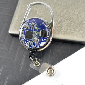 Puede incluir: Un portagafete retráctil plateado con un diseño de placa de circuito azul y plateado. El portagafete tiene un clip giratorio y una correa de plástico transparente.
