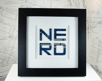 Op de afbeelding: Ingelijst kunstwerk met het woord "NERD" in blauwe printplaatletters. De letters staan tegen een witte achtergrond, omlijst door een zwart houten frame. Het kunstwerk wordt op een wit oppervlak weergegeven.