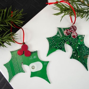 May include: Three green and red Christmas ornaments shaped like holly leaves made from recycled circuit boards. The ornaments have red berries and are strung with red ribbon.