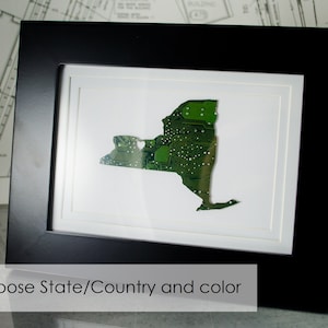 May include: A framed artwork depicting the outline of New York state, constructed from a green circuit board. The artwork is presented within a black frame, with a white mount. The text at the bottom reads "Choose State/Country and color".