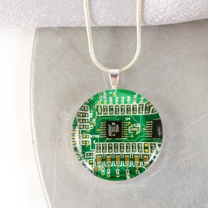 May include: A silver chain necklace with a round pendant featuring a green circuit board design. The pendant is encased in clear glass.