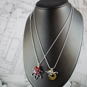 May include: Two silver necklaces with pendants on a black display stand. One pendant is a red and gold atom, the other is a silver and gold wolf.