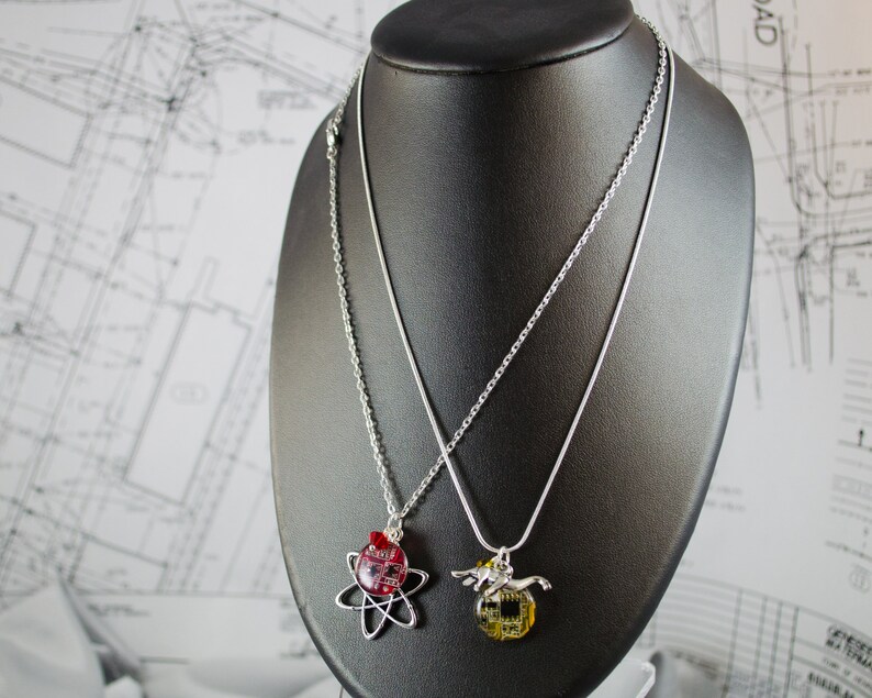 May include: Two silver necklaces with pendants on a black display stand. One pendant is a red and gold atom, the other is a silver and gold wolf.