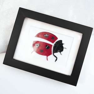 May include: A black framed picture featuring a red and black ladybug crafted from recycled circuit boards. The ladybug is depicted in a side profile with its wings extended.