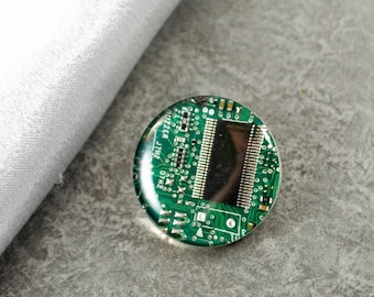 May include: A round, green and silver pin with a circuit board design. The circuit board has a black rectangular chip in the centre. The text "BUZZER JTIME 2H10" is printed on the circuit board.