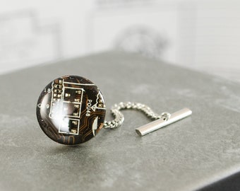 May include: A silver tie tack with a round, black and brown circuit board design. The circuit board has the text "POU48" printed on it.