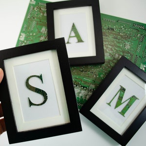 May include: Three black picture frames with white mats, each containing a letter of the alphabet, 'S', 'A', and 'M', made from green circuit boards.