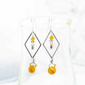 May include: A pair of silver earrings with a geometric diamond shape. Each earring features a small amber crystal, a silver bead, and a yellow ceramic capacitor. The earrings are hanging from silver hooks.