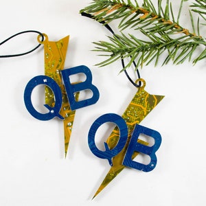 May include: Two handmade Christmas ornaments. Each ornament is shaped like a lightning bolt with the letters "QB" in blue. The lightning bolt is made from a circuit board with yellow and green details. A black string is attached for hanging.