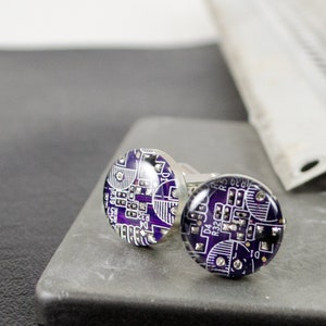 May include: A pair of round cufflinks featuring a circuit board design. The cufflinks have a dark purple background with white and silver circuit patterns. The cufflinks are made of metal and have a shiny, reflective surface.