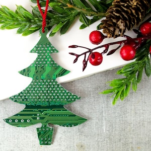 May include: A green Christmas tree ornament made from a circuit board, with a red string for hanging. The ornament is decorated with small silver dots and intricate circuit patterns. The image also includes a sprig of red berries and green foliage.