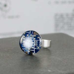 Puede incluir: Un anillo de plata con una incrustación circular azul y plateada que presenta un diseño de placa de circuito. La incrustación tiene texto que dice "R32 C51N R30 C51N R25".