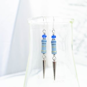 May include: A pair of silver earrings with blue and gold striped ceramic beads and blue crystal accents. The earrings have a pointed silver tip.