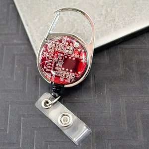 May include: A silver retractable badge holder with a red circuit board design. The holder has a swivel clip and a clear plastic strap.