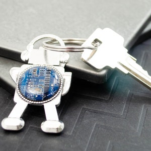 Puede incluir: Un llavero robot plateado con un diseño de placa de circuito azul. El llavero tiene un anillo de llave plateado.