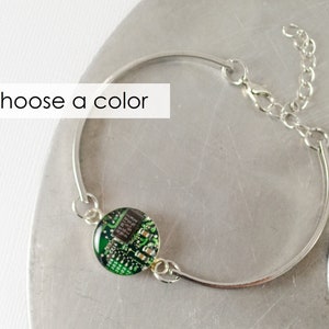 May include: A silver bangle bracelet with a green circuit board charm. The bracelet has a clasp closure and a chain extension.