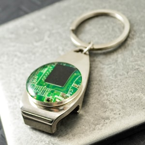 Puede incluir: Abrebotellas de llavero de plata con un diseño de placa de circuito verde. El llavero tiene un anillo para sujetarlo a las llaves.