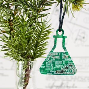 Könnte beinhalten: Ein grüner Weihnachtsbaumschmuck aus Leiterplatten in Form eines Bechers. Der Anhänger hängt an einer schwarzen Schnur und befindet sich vor einem grünen Kiefernzweig.