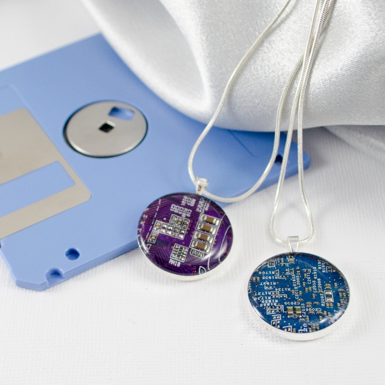 Puede incluir: Dos collares de plata con colgantes redondos que presentan dise&ntilde;os de placas de circuito p&uacute;rpura y azul. Los collares se muestran sobre un disquete azul y un fondo de tela blanca.