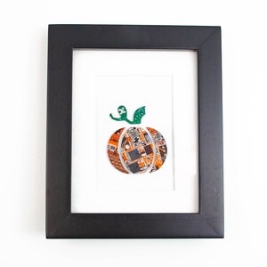 May include: A framed artwork featuring a pumpkin made from recycled circuit boards. The pumpkin is orange and brown with a green stem. The frame is black.
