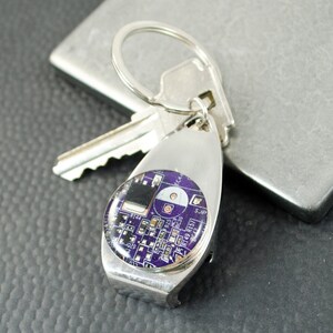 May include: A silver key ring with a purple circuit board design. The keyring has a bottle opener attached.