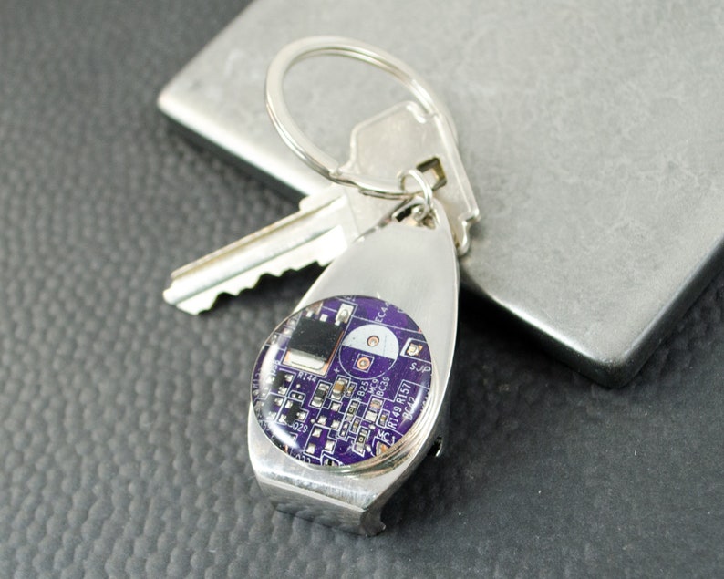 May include: A silver key ring with a purple circuit board design. The keyring has a bottle opener attached.