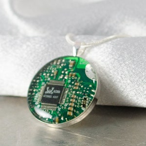 Puede incluir: Un collar de plata con un colgante redondo que presenta una placa de circuito verde encapsulada en resina. La placa de circuito tiene un peque&ntilde;o chip de circuito integrado con el texto "S196L 4230062 431F" impreso en &eacute;l.