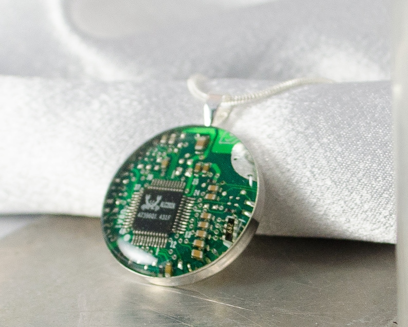 Puede incluir: Un collar de plata con un colgante redondo que presenta una placa de circuito verde encapsulada en resina. La placa de circuito tiene un peque&ntilde;o chip de circuito integrado con el texto "S196L 4230062 431F" impreso en &eacute;l.