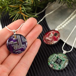 Puede incluir: Tres collares de plata con colgantes redondos que muestran placas de circuito de diferentes colores. Los colgantes son morado, rojo y verde.