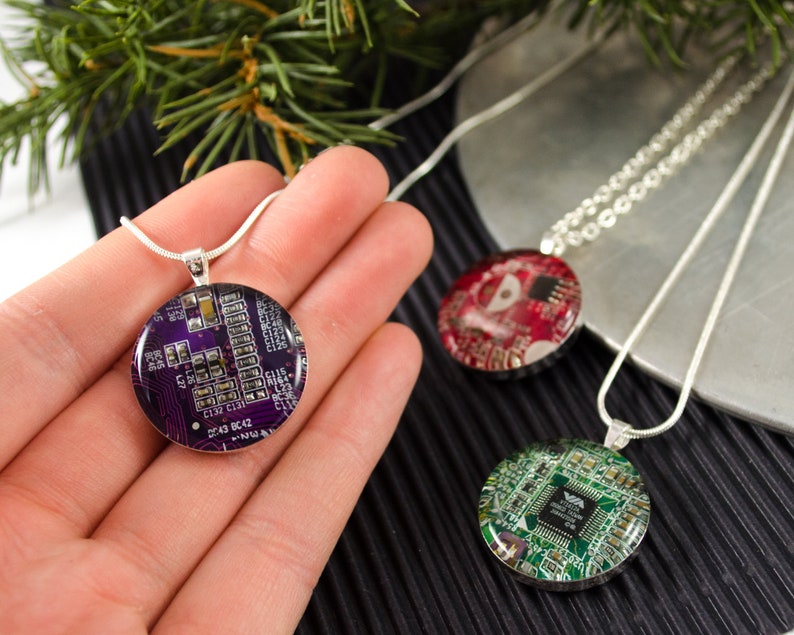 Puede incluir: Tres collares de plata con colgantes redondos que muestran placas de circuito de diferentes colores. Los colgantes son morado, rojo y verde.