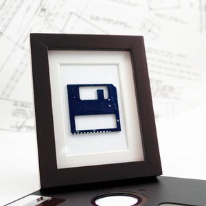 May include: A framed artwork featuring a dark blue circuit board in a black frame with a white mat. The artwork is displayed on a white surface with a floppy disk and blueprints in the background. The frame is rectangular.