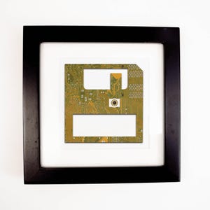 May include: Framed artwork featuring a vintage computer floppy disk. The disk's circuit board is visible, showcasing green and gold tones. The artwork is set against a white background within a black frame, creating a modern aesthetic.