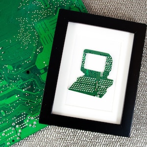 May include: A black picture frame with a white mount and a green circuit board cutout inside. The circuit board is a common component found in computers and other electronic devices.