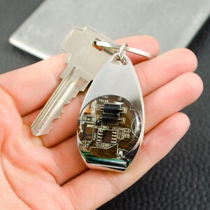 May include: A silver keyring with a clear epoxy resin circle containing a circuit board. The circuit board has black components and the text "PO146" is visible.