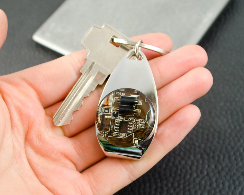 May include: A silver keyring with a clear epoxy resin circle containing a circuit board. The circuit board has black components and the text "PO146" is visible.