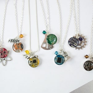 May include: Seven silver necklaces with different charms. Each charm features a circuit board design with a different colored crystal. The charms include a beaker, a microscope, a gear, a robot, an atom, and the word "NERD".