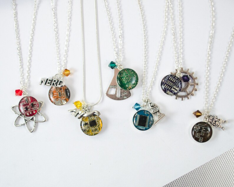 May include: Seven silver necklaces with different charms. Each charm features a circuit board design with a different colored crystal. The charms include a beaker, a microscope, a gear, a robot, an atom, and the word "NERD".