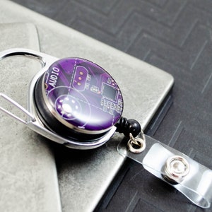 Puede incluir: Un carrete de identificación retráctil plateado con un diseño de placa de circuito morado y negro. El carrete tiene un clip plateado y una correa de plástico transparente.